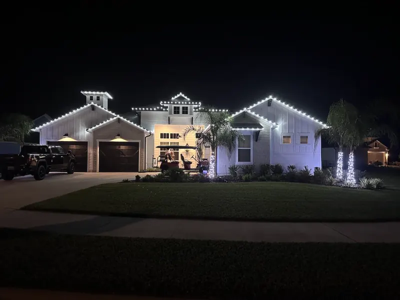 Modern farmhouse at night with bright white lights along the roof edges and lit palm trunks
