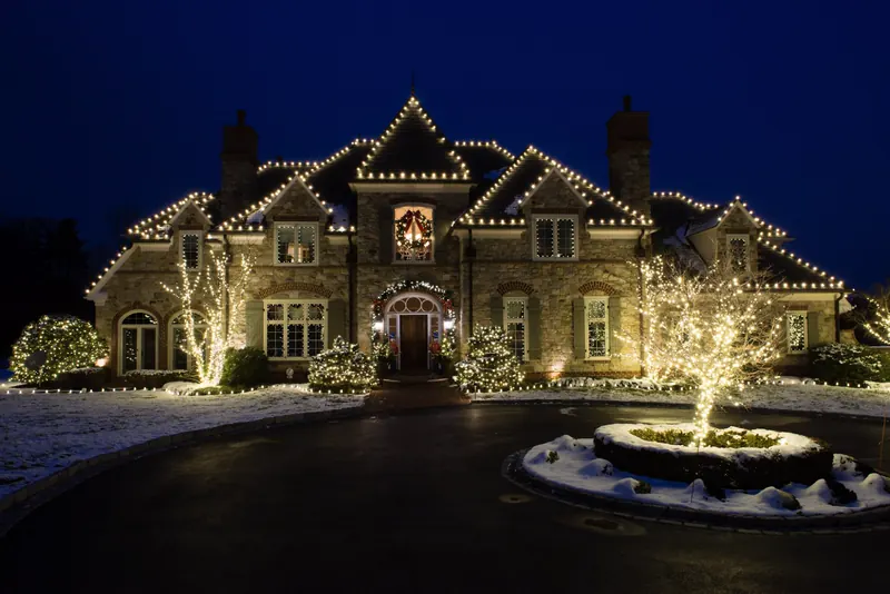 Stone manor with warm white lights on every roof edge, wreaths at the windows and snow on the lawn